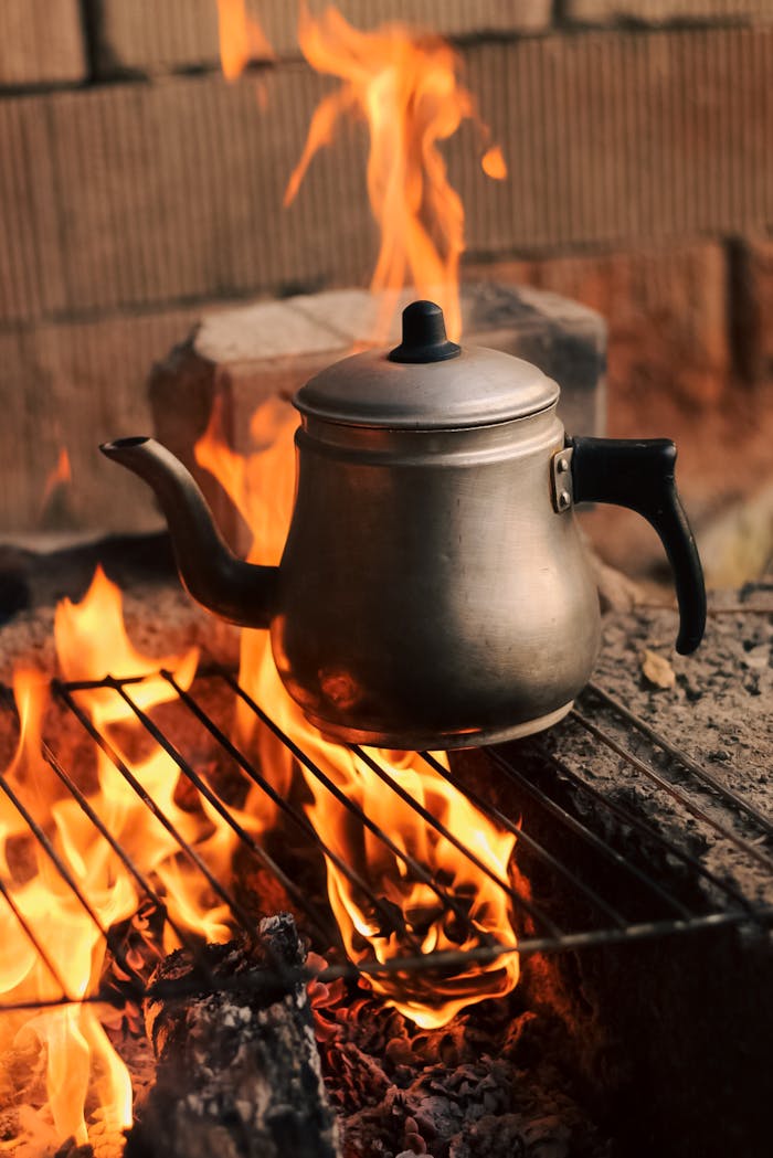 A classic metal kettle heats over an open flame on a grill, set outdoors for rustic cooking.